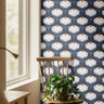Chantelune Noir 2249 navy blue floral wallpaper featuring repeating white coneflowers with warm golden centers, styled in a cozy room with a natural wood chair, trailing green plant, and sunlit window.