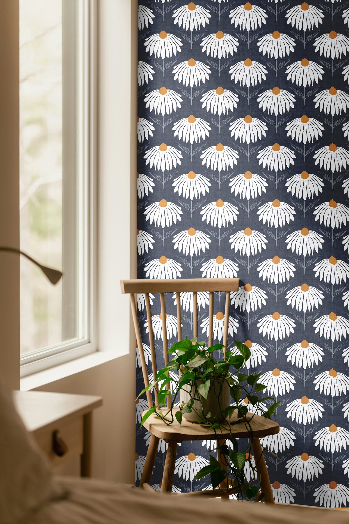 Chantelune Noir 2249 navy blue floral wallpaper featuring repeating white coneflowers with warm golden centers, styled in a cozy room with a natural wood chair, trailing green plant, and sunlit window.