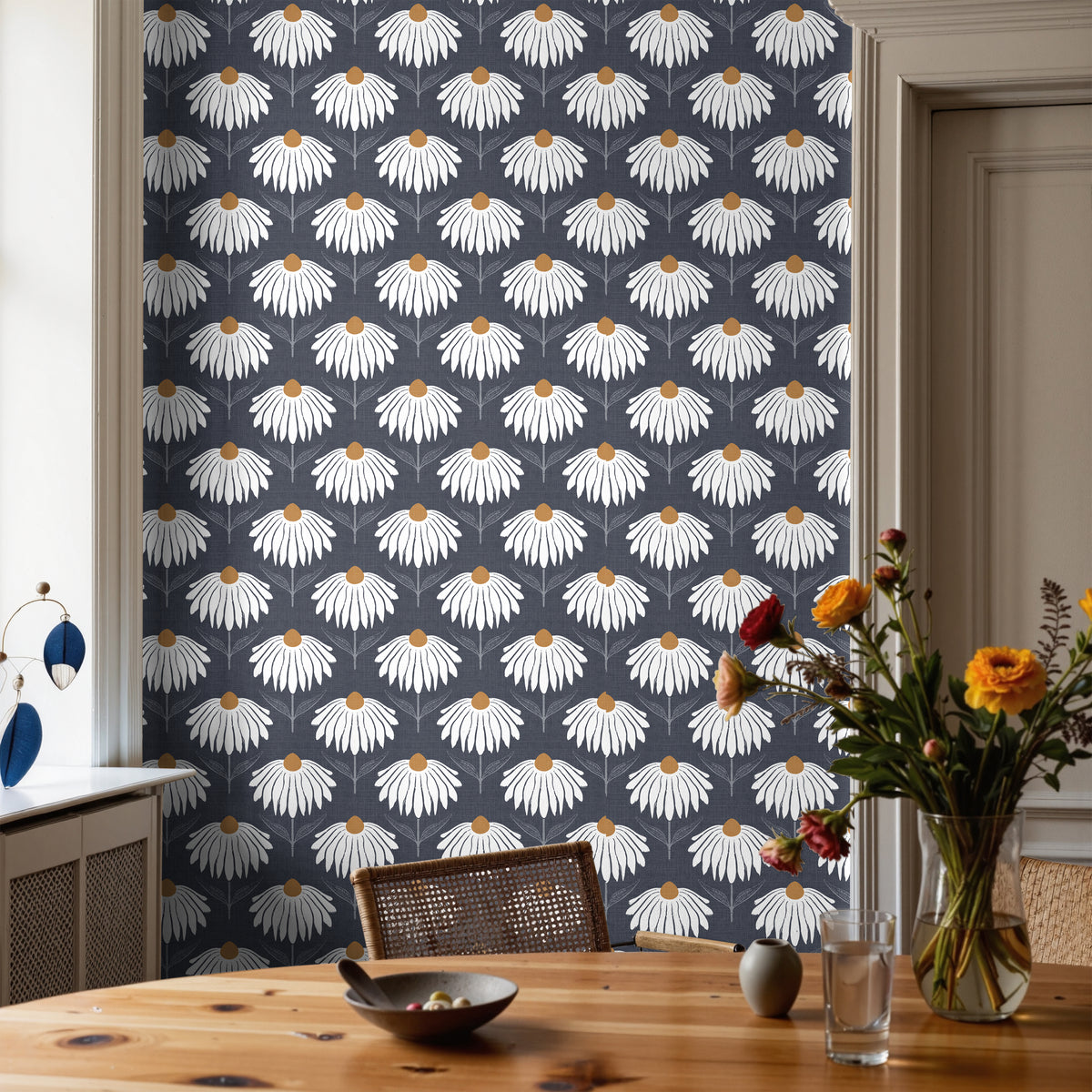 Chantelune Noir 2249 navy blue floral wallpaper featuring repeating white coneflowers with warm golden centers, styled in a dining room with a natural wood table, woven chairs, and a colorful floral arrangement.