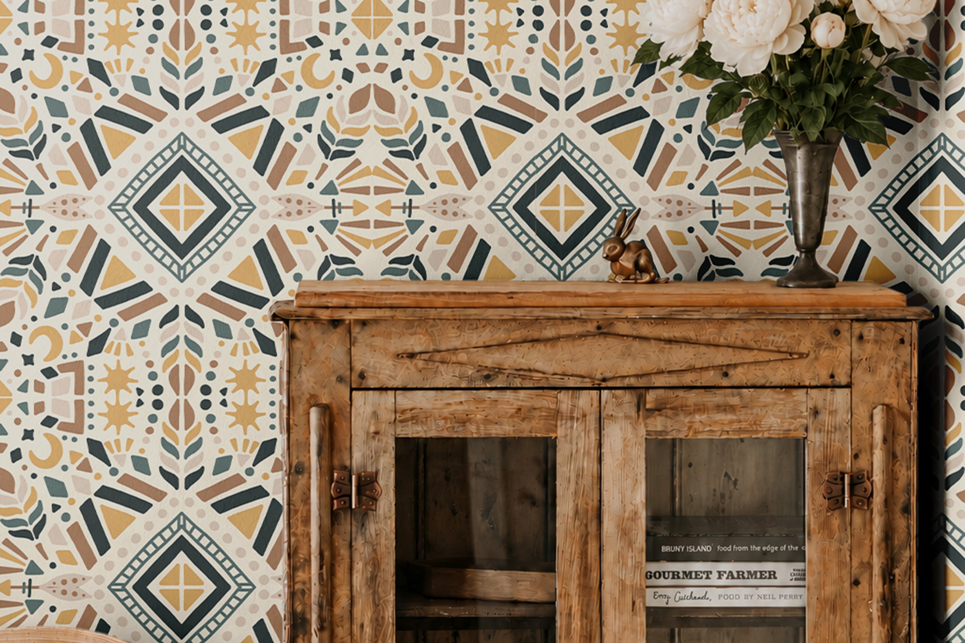 Geometric Boho 2377 wallpaper featuring a colorful bohemian geometric pattern in mustard yellow, muted teal, terracotta, and cream, styled in a dining room with a rustic wooden cabinet and white flowers.