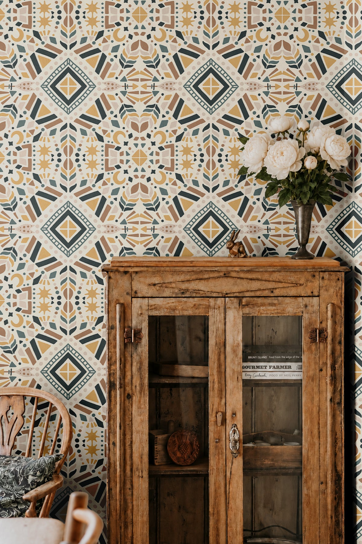 Geometric Boho 2377 wallpaper featuring a colorful bohemian geometric pattern in mustard yellow, muted teal, terracotta, and cream, styled in a dining room with a rustic wooden cabinet and white flowers.