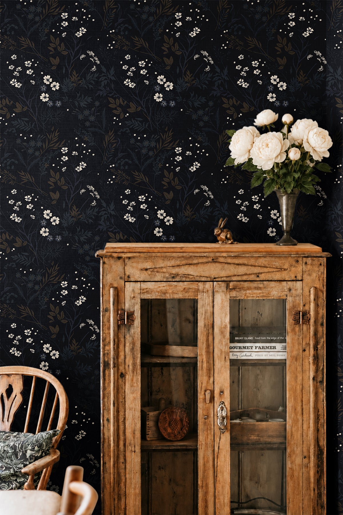 ink and elderflower 3552 moody floral wallpaper behind a rustic wood cabinet with white flowers in a vase and a wood chair