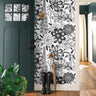 Monochrome Meadow 764 black and white botanical wallpaper featuring oversized vintage wildflowers and detailed floral illustrations, styled in a modern entryway with a dark green door, natural wood flooring, black rain boots, and an umbrella.