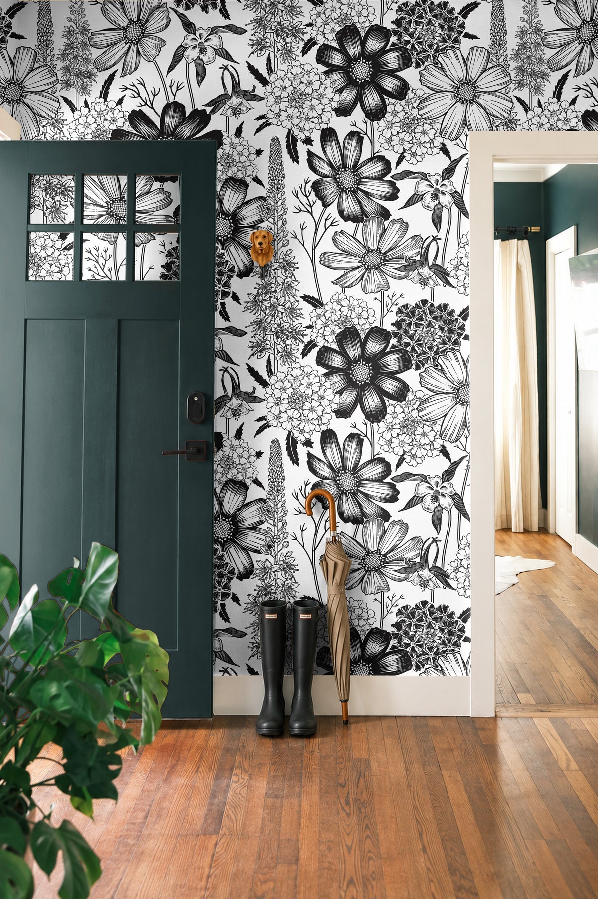 Monochrome Meadow 764 black and white botanical wallpaper featuring oversized vintage wildflowers and detailed floral illustrations, styled in a modern entryway with a dark green door, natural wood flooring, black rain boots, and an umbrella.